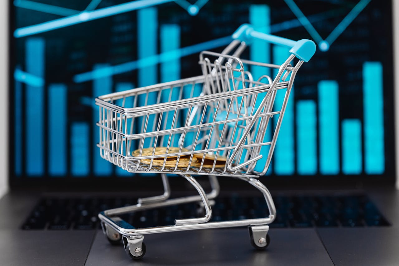 about-01 A miniature shopping cart with coins sits on a laptop displaying financial graphs, symbolizing e-commerce and online shopping trends.
